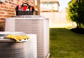 Bag of repairman's work tools, gloves on top of air conditioner units outside a brick home.  Service industry, working class.